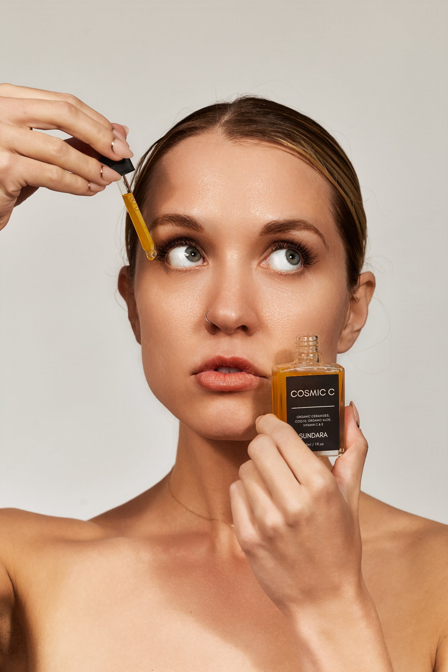Woman applying a dropper of cosmetic oil to her face with a neutral background