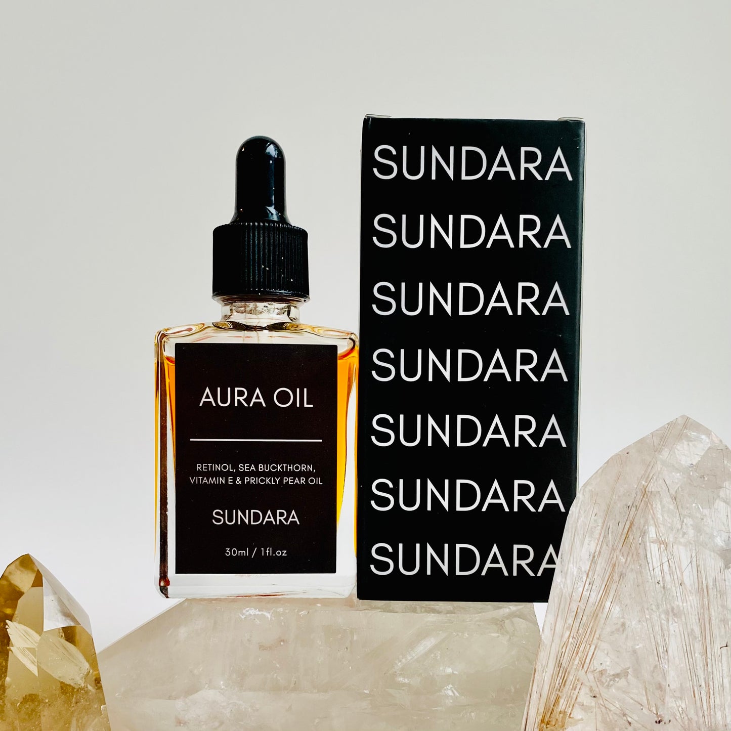 Two bottles of Aura Oil serum with dropper caps, one standing upright and the other lying in front, against a white background with a large crystal next to them. Retinol, sea buckthorn, vitamin e, prickle pear oil