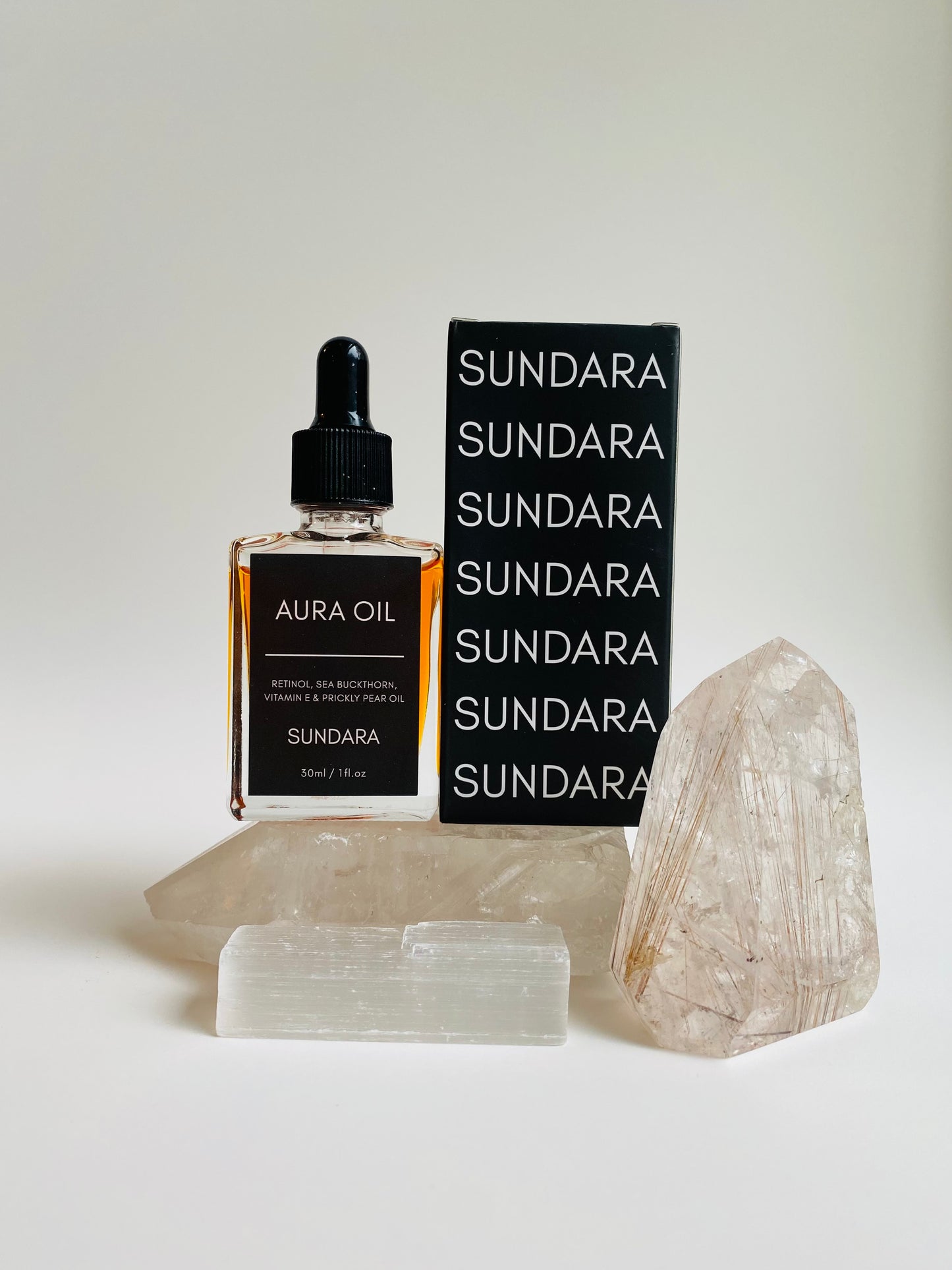 Two bottles of Aura Oil serum with dropper caps, one standing upright and the other lying in front, against a white background with a large crystal next to them. Retinol, sea buckthorn, vitamin e, prickle pear oil