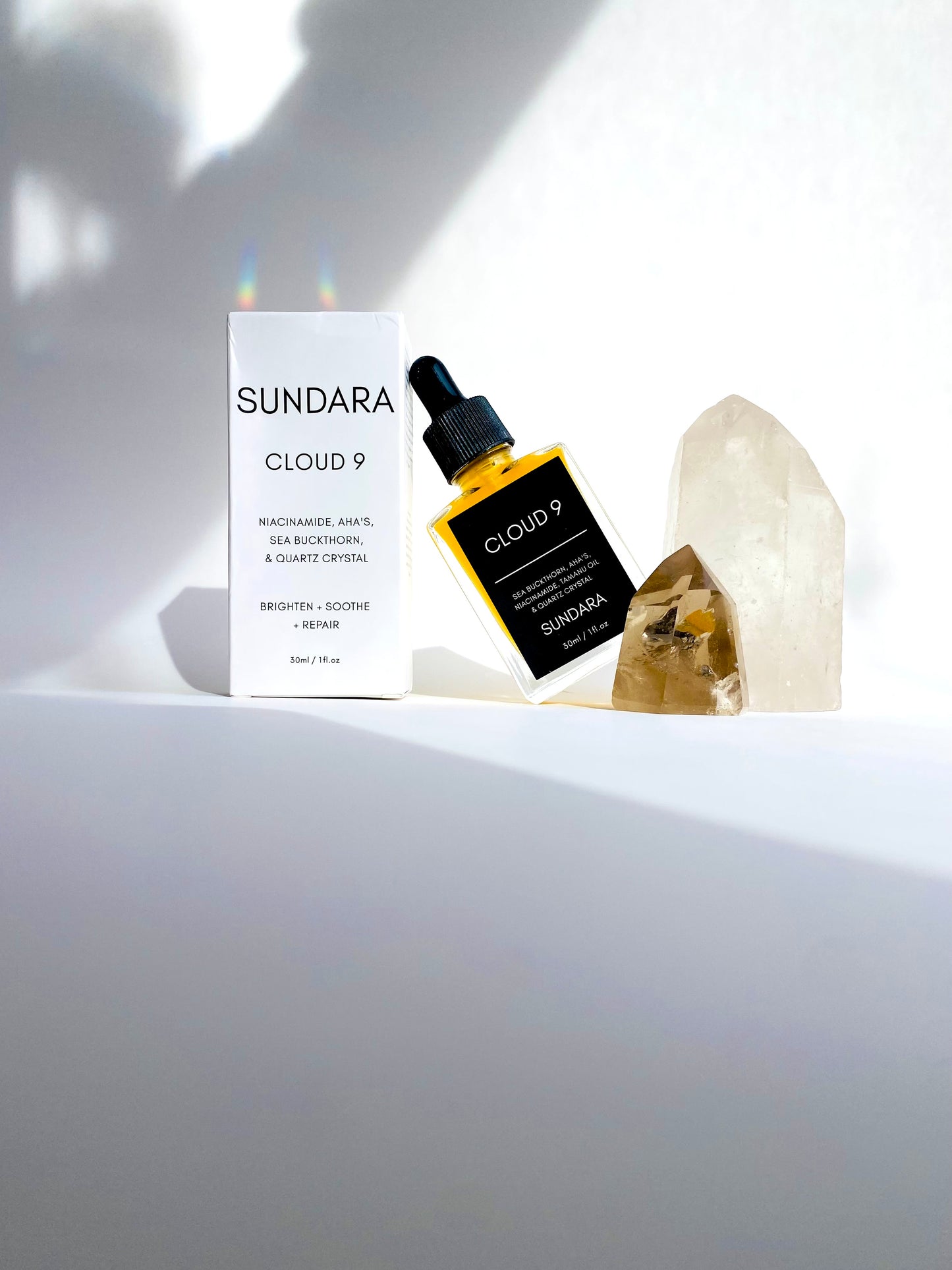 A bottle of 'Cloud 9' skin serum by Sundara, with a sea buckthorn oil dropper, placed on a surface with a crystal. Niacinimide, AHA's, Sea Buckthorn, Quartz Crystal for brightening, soothing, and repairing the skin barrier as an all natural, plant based, skincare serum.