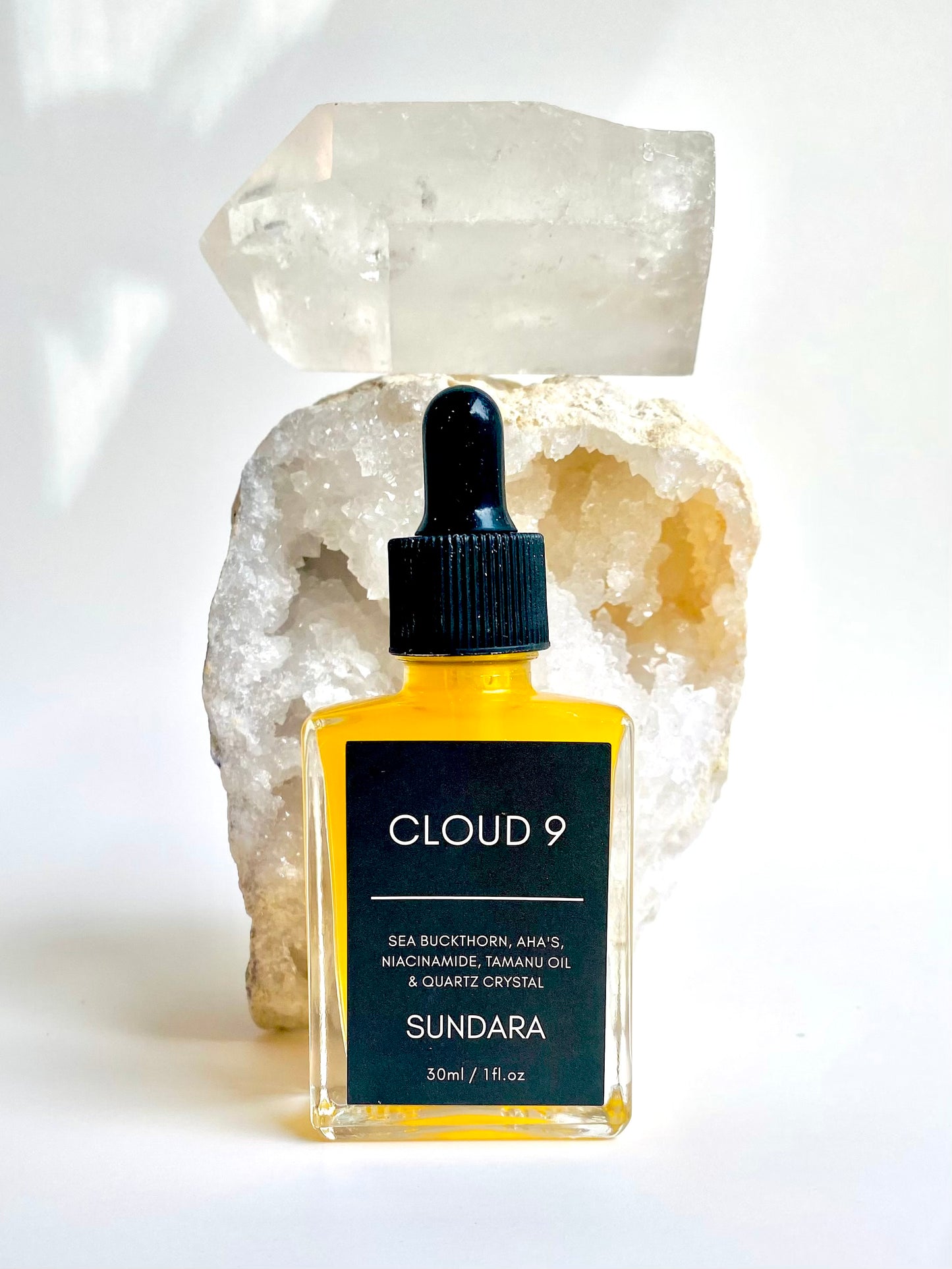 A bottle of 'Cloud 9' skin serum by Sundara, with a sea buckthorn oil dropper, placed on a surface with a crystal. Niacinimide, AHA's, Sea Buckthorn, Quartz Crystal for brightening, soothing, and repairing the skin barrier as an all natural, plant based, skincare serum.