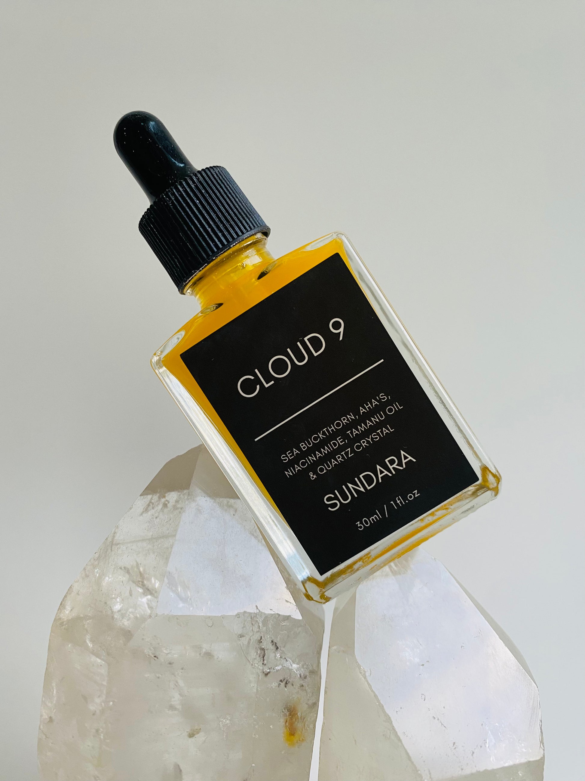 A bottle of 'Cloud 9' skin serum by Sundara, with a sea buckthorn oil dropper, placed on a surface with a crystal. Niacinimide, AHA's, Sea Buckthorn, Quartz Crystal for brightening, soothing, and repairing the skin barrier as an all natural, plant based, skincare serum.