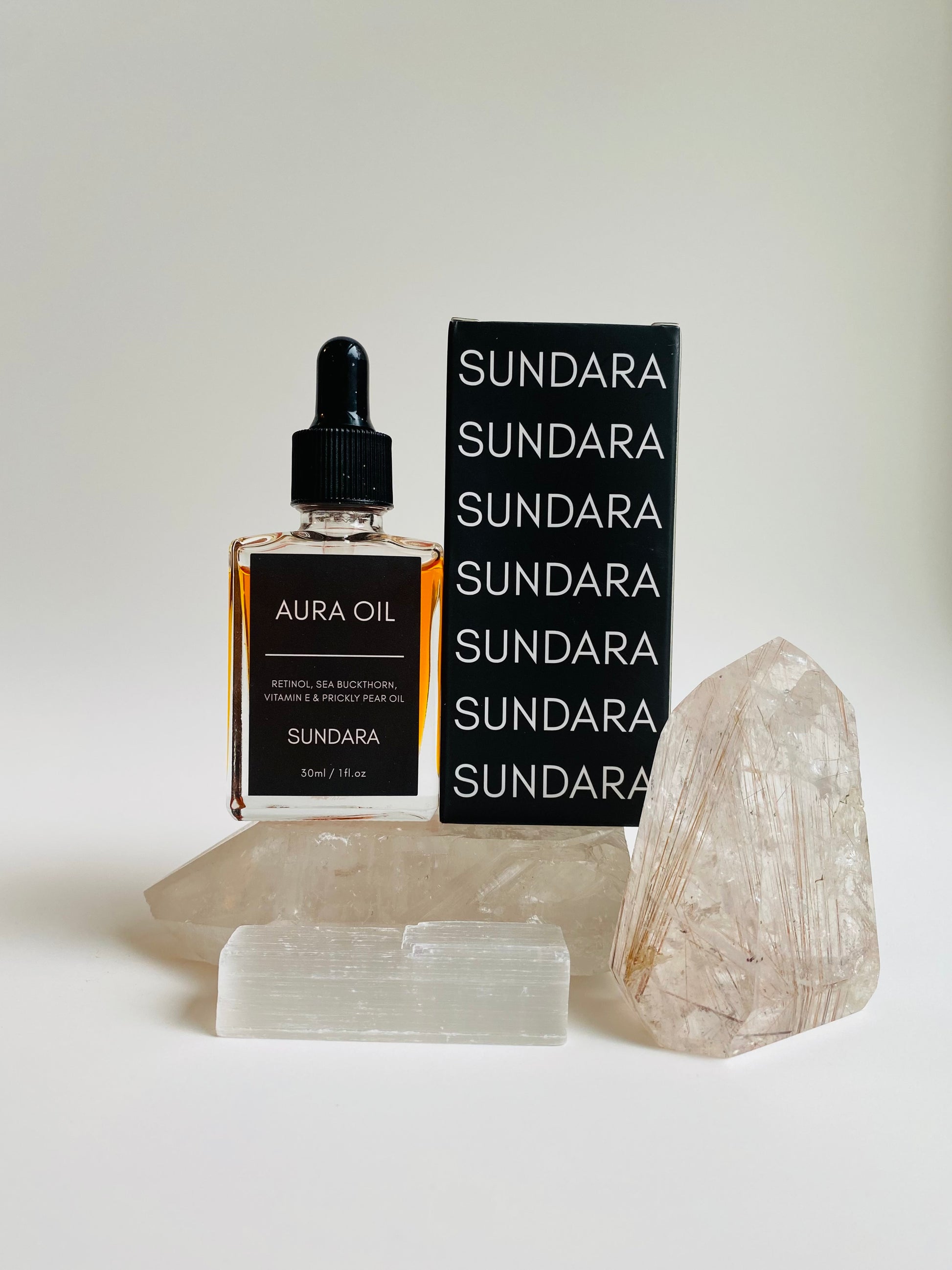Two bottles of Aura Oil serum with dropper caps, one standing upright and the other lying in front, against a white background with a large crystal next to them. Retinol, sea buckthorn, vitamin e, prickle pear oil