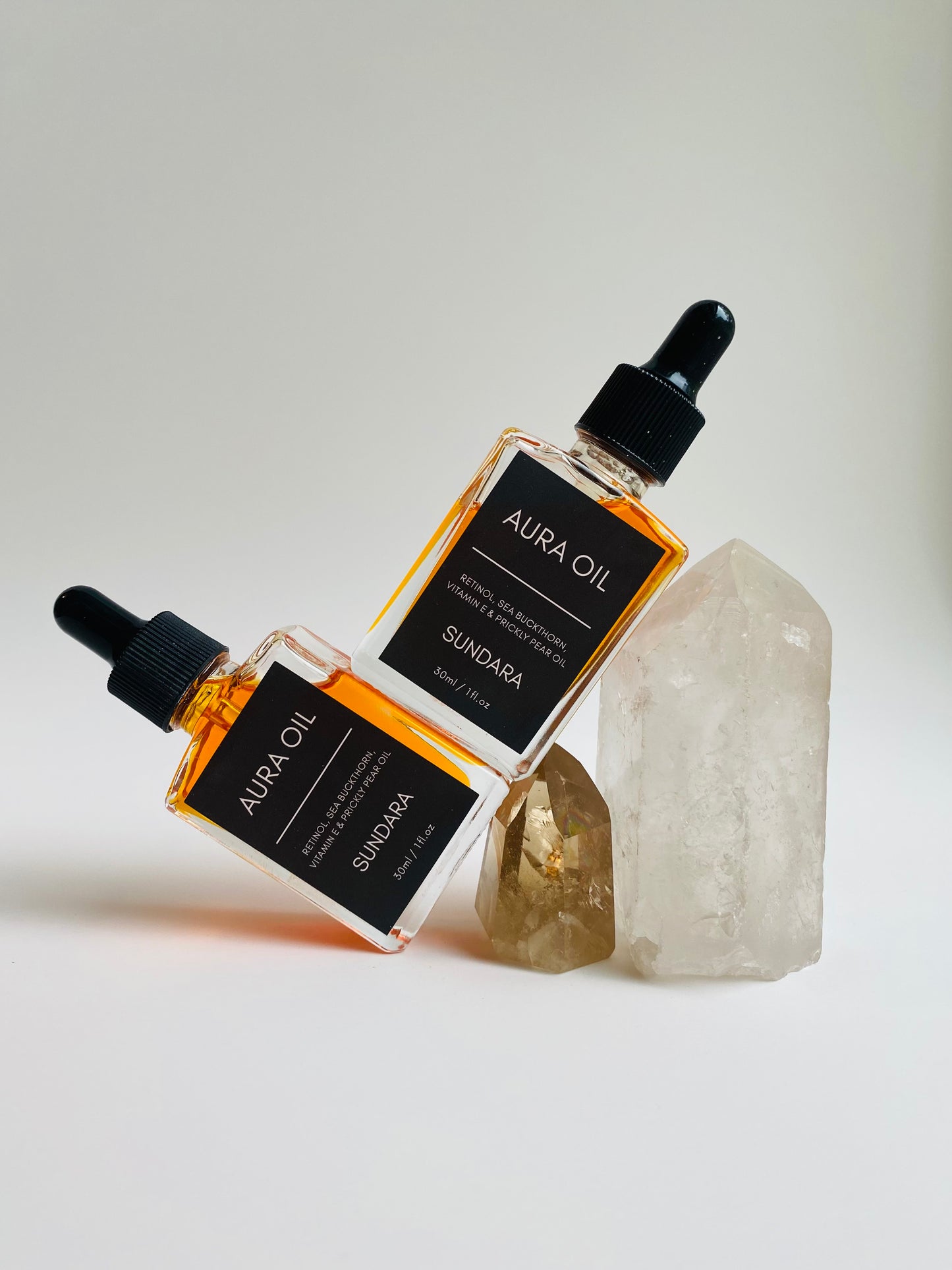 Two bottles of Aura Oil serum with dropper caps, one standing upright and the other lying in front, against a white background with a large crystal next to them. Retinol, sea buckthorn, vitamin e, prickle pear oil