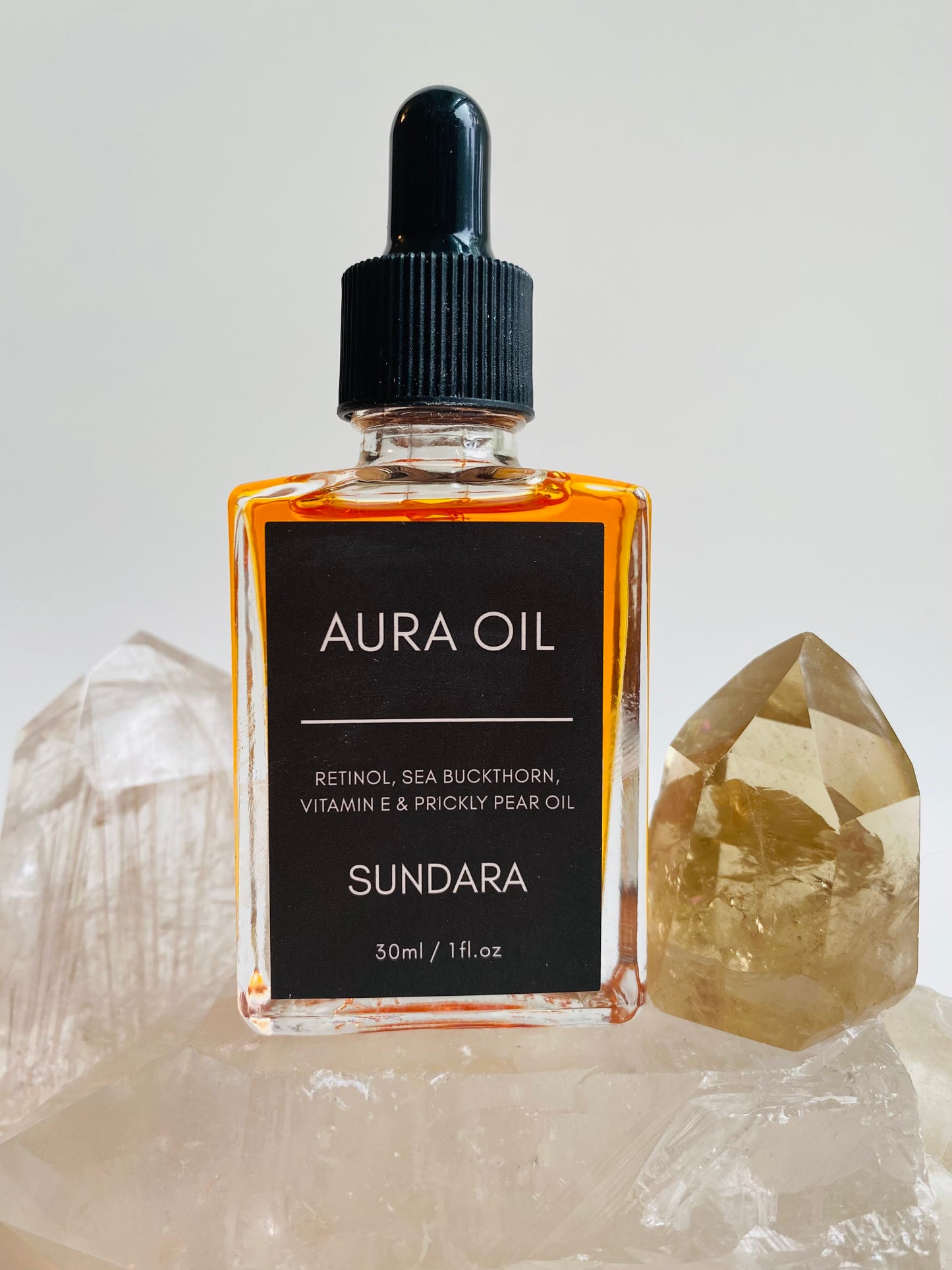 Two bottles of Aura Oil serum with dropper caps, one standing upright and the other lying in front, against a white background with a large crystal next to them. Retinol, sea buckthorn, vitamin e, prickle pear oil